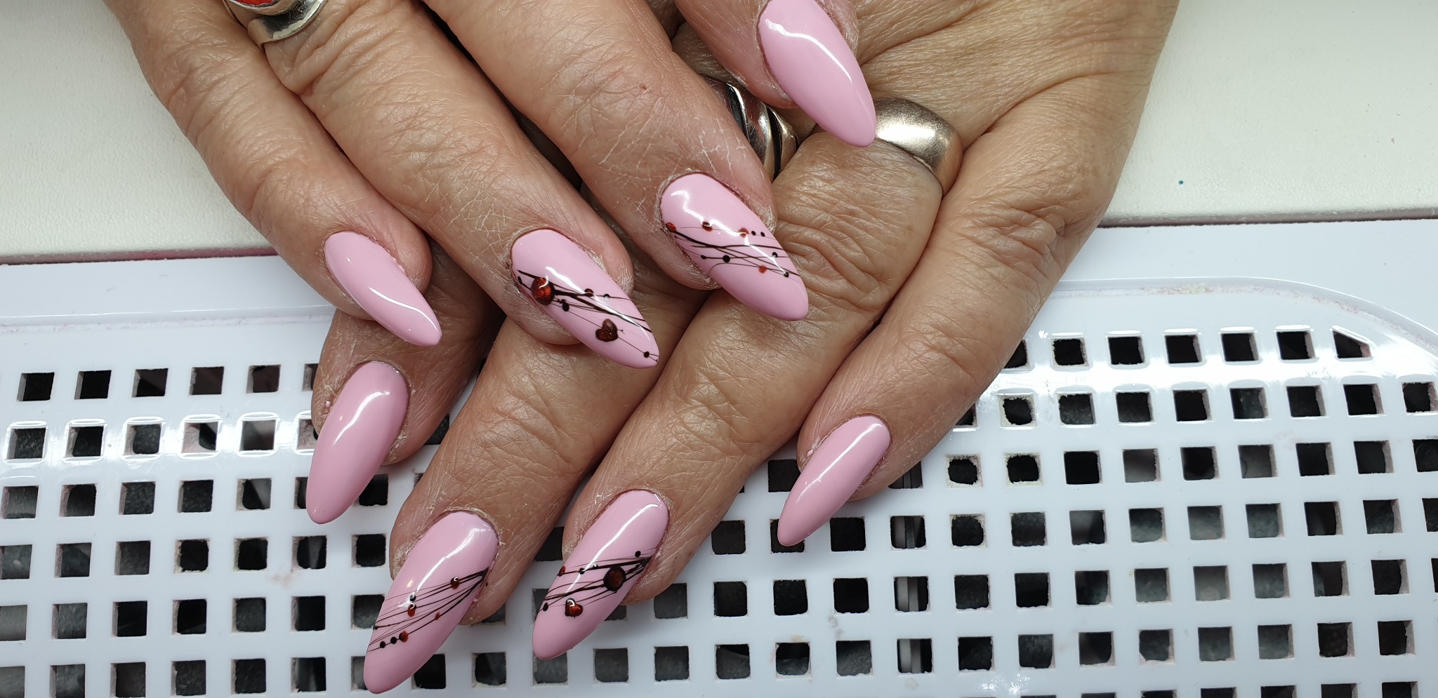 Dłonie z długimi, migdałowymi paznokciami pomalowanymi na jasny róż z delikatnym zdobieniem w postaci cienkich, czarnych linii i małych, czerwonych kropek.