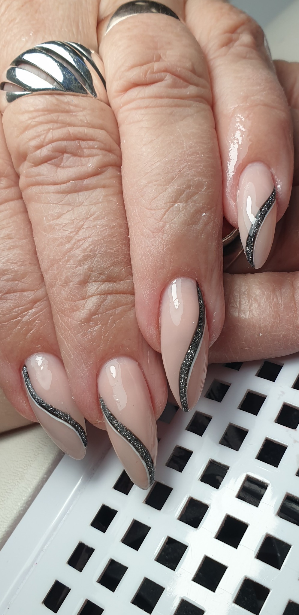 Dłoń z beżowymi paznokciami zdobionymi falowaną srebrną linią brokatu, widoczny srebrny pierścionek na palcu.