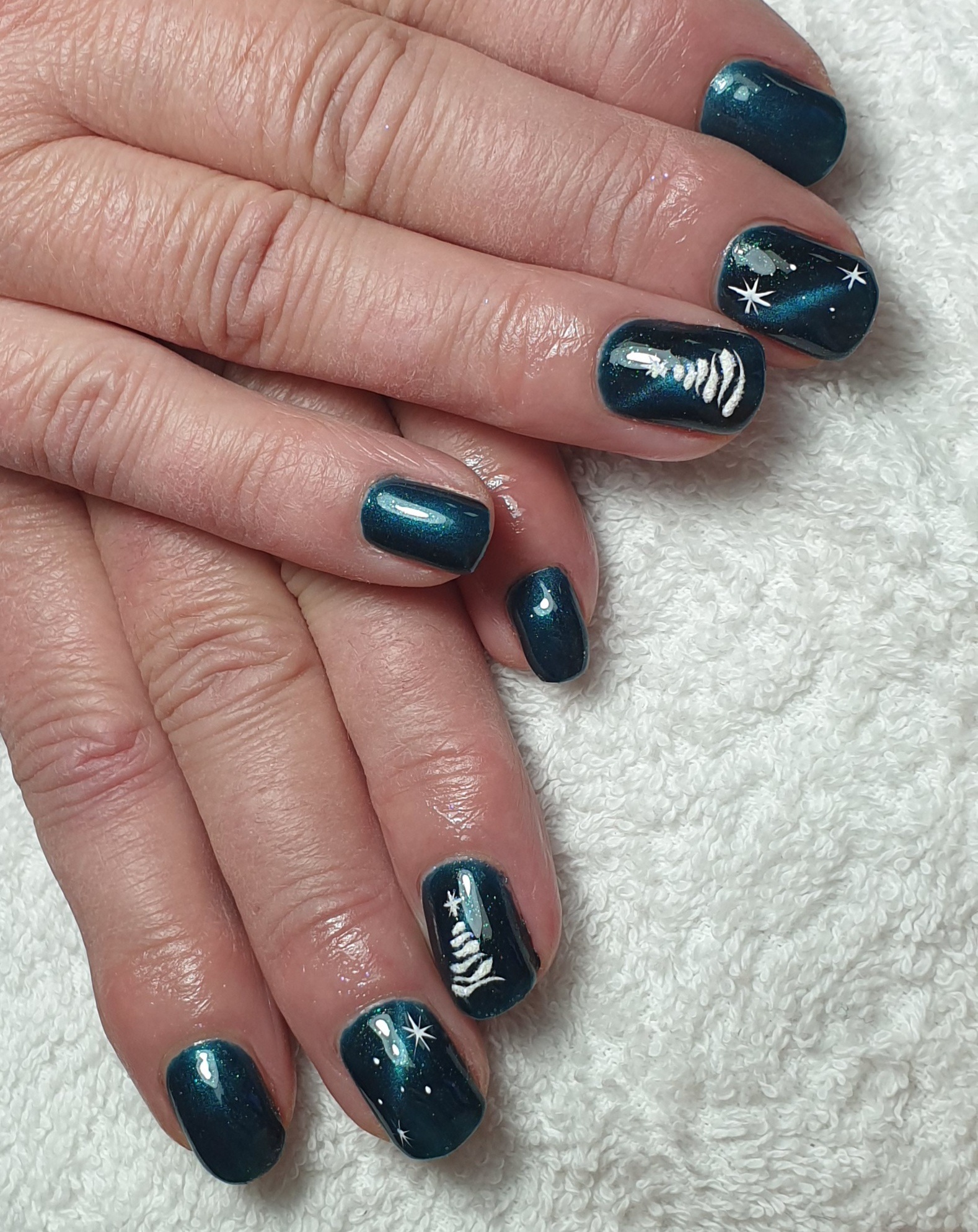 Dłonie z ciemnozielonymi paznokciami, na których widnieją białe, świąteczne wzory: choinka i gwiazdki, na tle białego ręcznika.