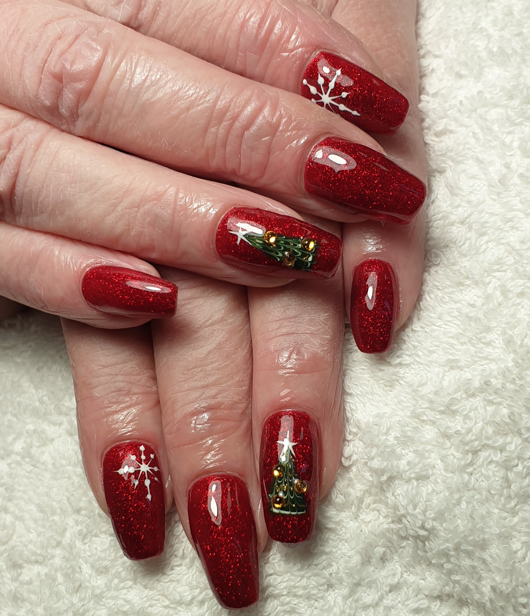 Czerwone, brokatowe paznokcie z motywami świątecznymi: białe płatki śniegu i choinki z ozdobami, na tle białego ręcznika.