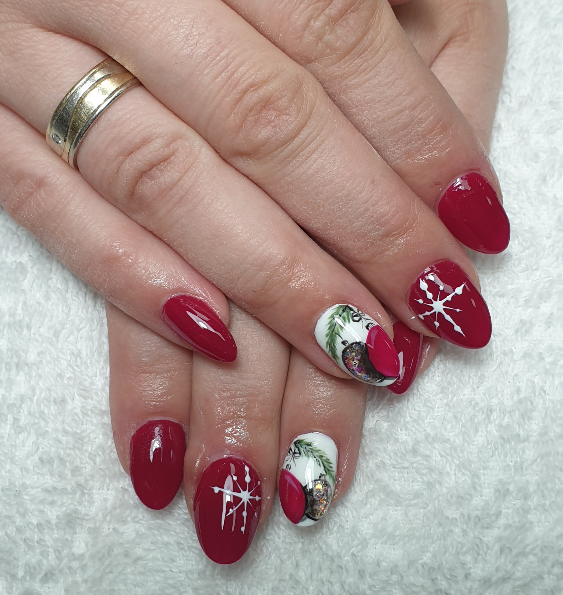 Dłonie z czerwonymi paznokciami w kształcie migdała, ozdobionymi białymi śnieżynkami i motywami świątecznymi, w tym bombkami i gałązkami choinki, na tle białego ręcznika.