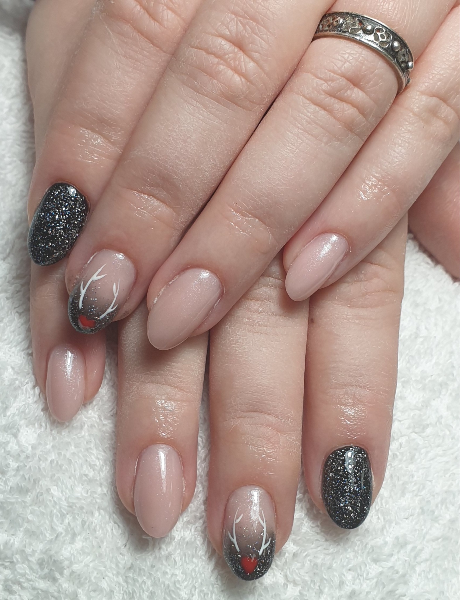 Dłonie z owalnymi paznokciami: dwa w kolorze nude, dwa pokryte czarnym brokatem i jeden z wzorem renifera z czerwonym nosem i białymi rogami na szaro-czarnym tle.