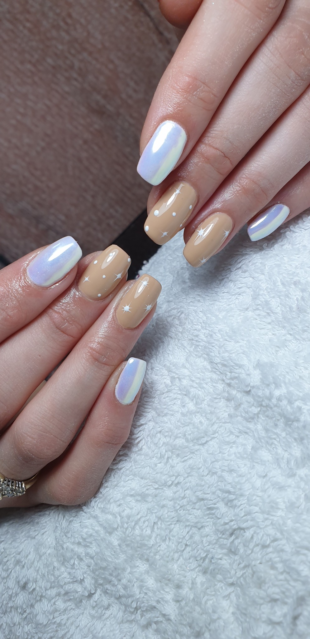 Dłonie z kwadratowymi paznokciami, jeden z efektem lustra, a drugi beżowy z białymi kropkami i gwiazdkami, na białym, puszystym ręczniku.