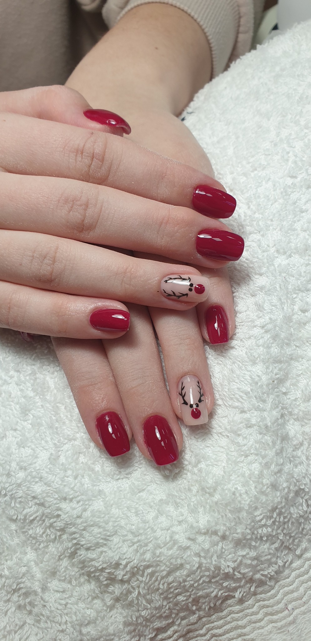 Dłonie z czerwonymi paznokciami i świątecznym zdobieniem renifera na dwóch paznokciach, ułożone na białym ręczniku.