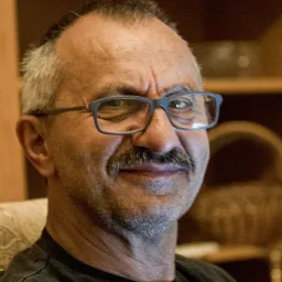 Portret uśmiechniętego mężczyzny w okularach z szarą brodą i wąsami, na tle drewnianego regału.