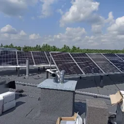 Panele słoneczne zamontowane na płaskim dachu, widoczne elementy wentylacji i opakowania po komponentach, w tle las i błękitne niebo z chmurami.