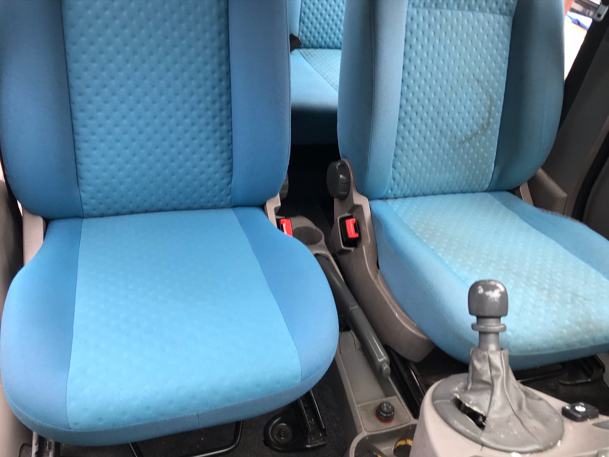 Wnętrze samochodu z jasnoniebieską tapicerką foteli, widoczne ślady użytkowania na siedzeniu pasażera oraz uszkodzona osłona dźwigni zmiany biegów.