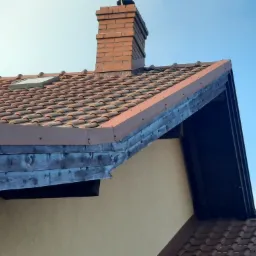 Ceglany komin na dachu z brązowej dachówki ceramicznej, widoczne okno dachowe i drewniane elementy wykończeniowe w kolorze ciemnego brązu.