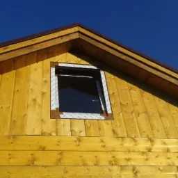 Fragment ściany zewnętrznej domu z desek w odcieniach żółci z uchylonym oknem na poddaszu, widoczny brązowy okap dachu na tle błękitnego nieba.