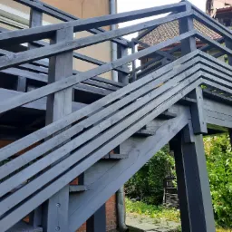 Drewniane schody tarasowe pomalowane na szaro, prowadzące na podwyższony taras, z widocznymi elementami konstrukcyjnymi i roślinnością w tle.