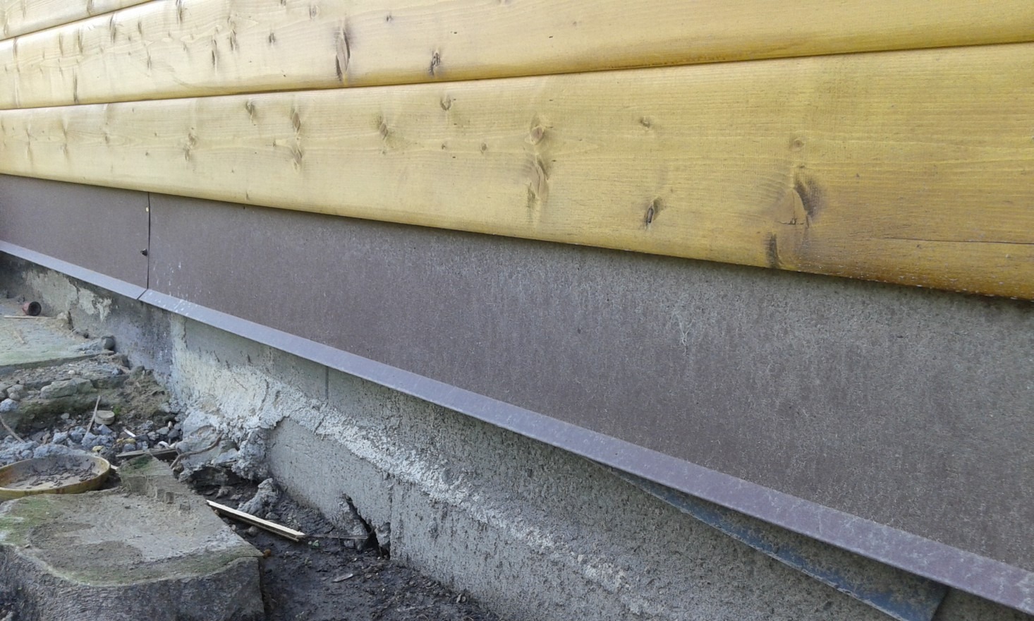 Fragment ściany zewnętrznej budynku z drewnianą boazerią i szarą, metalową listwą przypodłogową oraz betonową podmurówką w trakcie prac wykończeniowych.