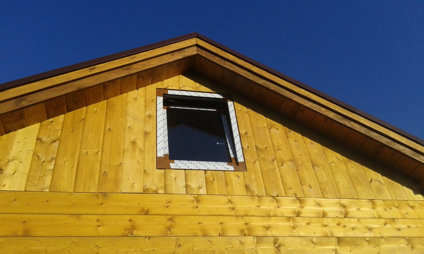 Fragment ściany zewnętrznej domu z desek w odcieniach żółci z uchylonym oknem na poddaszu, widoczny brązowy okap dachu na tle błękitnego nieba.