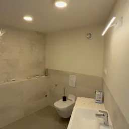 Jasna łazienka po remoncie z beżowymi płytkami, umywalką na blacie imitującym marmur, toaletą podwieszaną i oświetleniem punktowym. Widoczny fragment opakowania.