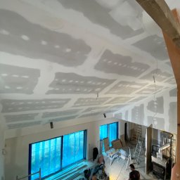 ProDremont Dmytro Nimenko - Sufit z płyt kartonowo-gipsowych w trakcie montażu, widok z góry. Widoczne łączenia płyt i fragmenty konstrukcji drewnianej. W tle okna z niebieską folią ochronną.