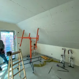 Montaż płyt na poddaszu: widok na skośny sufit z zamontowanymi płytami, podnośnik do płyt, drabina, narzędzia leżące na podłodze, fragment okna i sylwetka pracownika w lewym dolnym rogu.