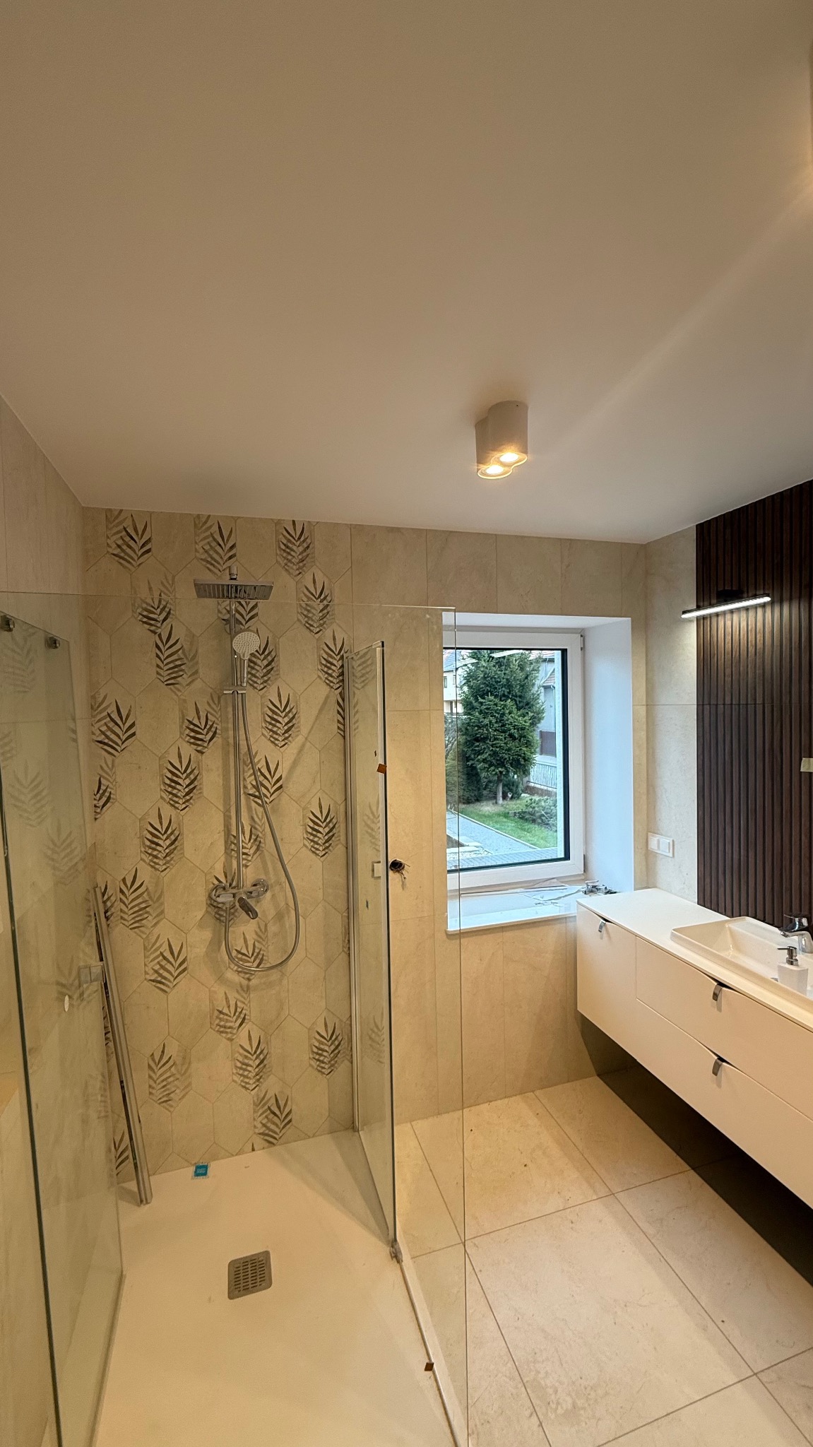 Nowoczesna łazienka z kabiną prysznicową typu walk-in, beżowymi heksagonalnymi płytkami z motywem liści i oknem z widokiem na zieleń.