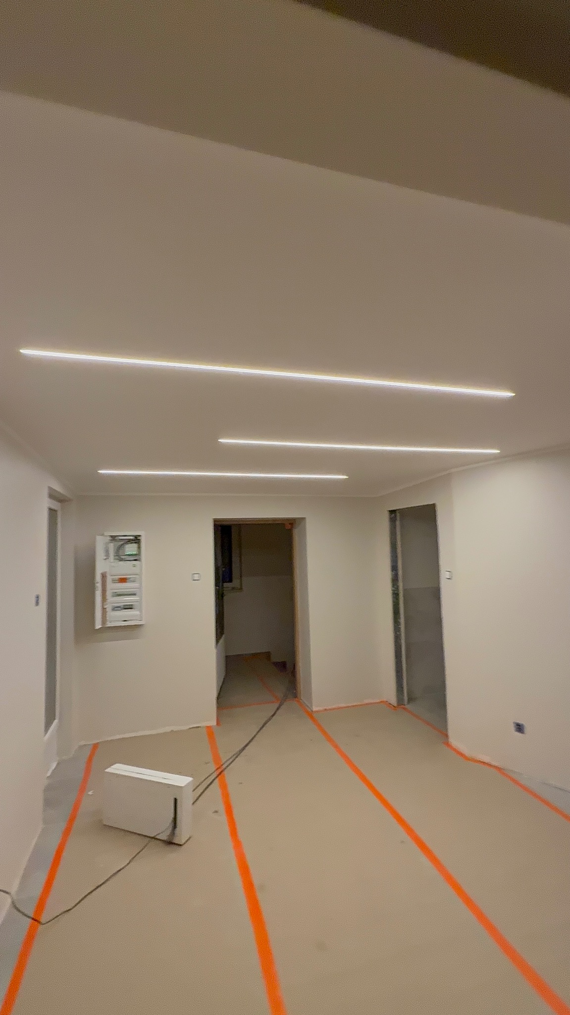 Wnętrze w trakcie remontu z nowym oświetleniem liniowym LED na suficie i zabezpieczoną podłogą. Widoczna skrzynka elektryczna na ścianie i przygotowane przejścia między pomieszczeniami.