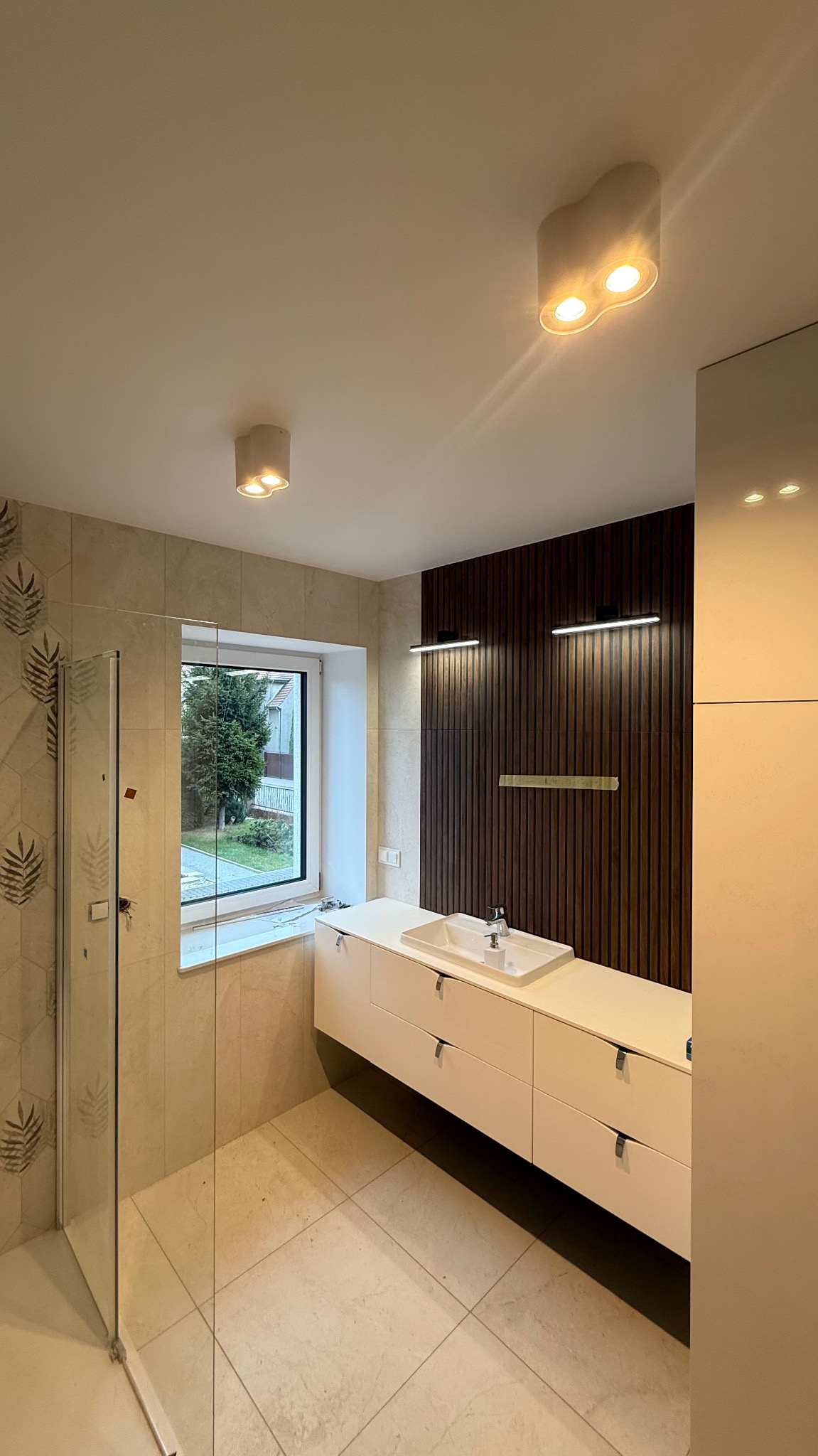 Nowoczesna łazienka z kabiną prysznicową, beżowymi płytkami, długą białą szafką pod umywalką i ścianą z drewnianych paneli. Widoczne okno i oświetlenie sufitowe.