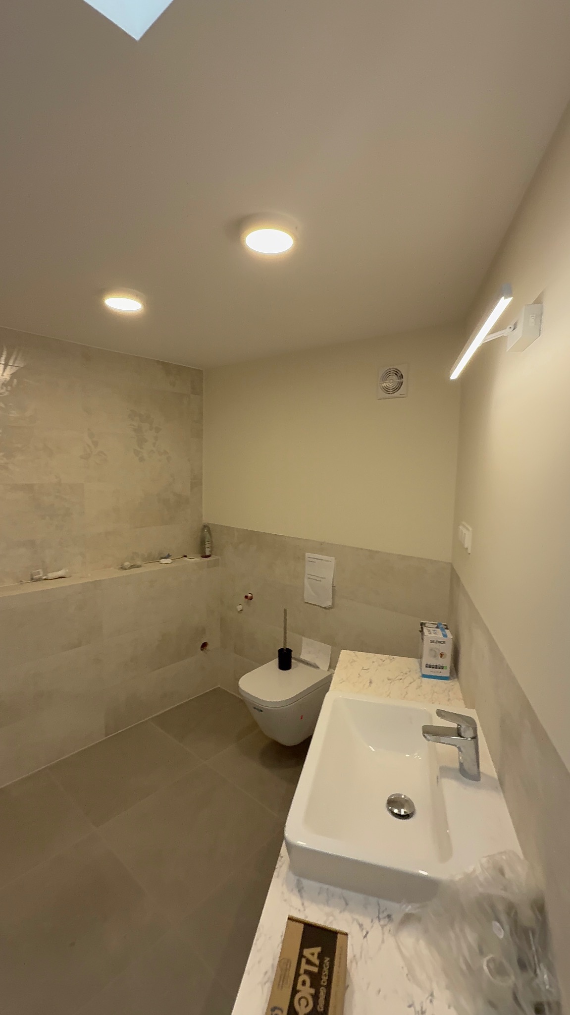 Jasna łazienka po remoncie z beżowymi płytkami, umywalką na blacie imitującym marmur, toaletą podwieszaną i oświetleniem punktowym. Widoczny fragment opakowania.