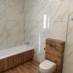 Wykończona łazienka z wanną obudowaną drewnem, wiszącą toaletą z obudową z drewnianych paneli, podłogą z paneli imitujących drewno i ścianami wyłożonymi płytkami imitującymi marmur.