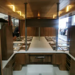 Wnętrze mobilnej kuchni z drewnianymi szafkami, białym blatem i chromowanymi pojemnikami na żywność, oświetlone punktowymi lampami sufitowymi.