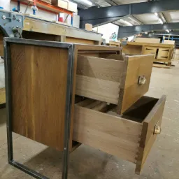 Drewniana szafka z dwoma wysuniętymi szufladami, metalowa rama, widoczne połączenia stolarskie typu jaskółczy ogon, w tle warsztat stolarski.