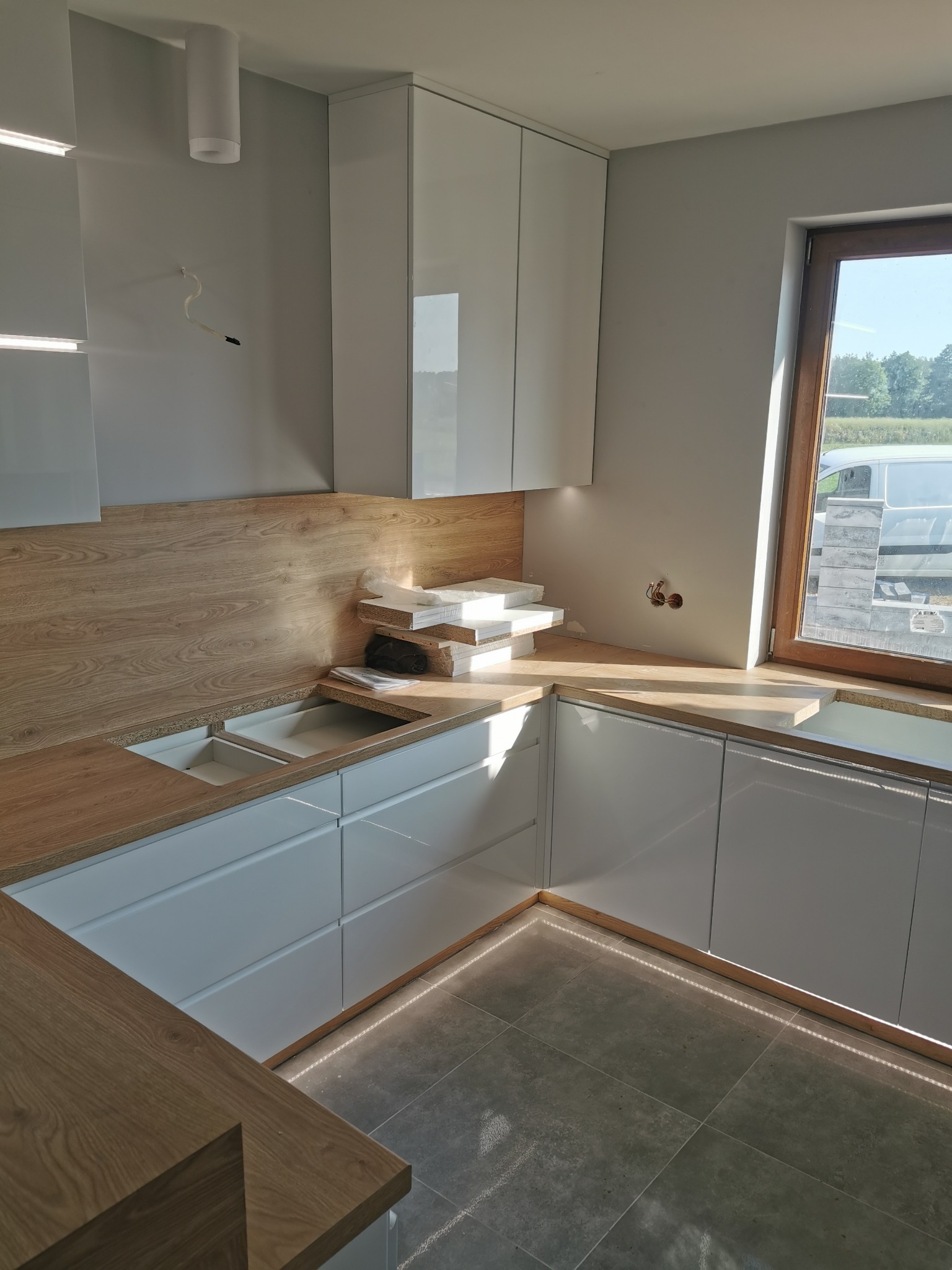 Montaż białych szafek kuchennych z drewnianym blatem, widoczne niedokończone podłączenia elektryczne i brakujące elementy zlewu, oświetlenie podszafkowe LED.