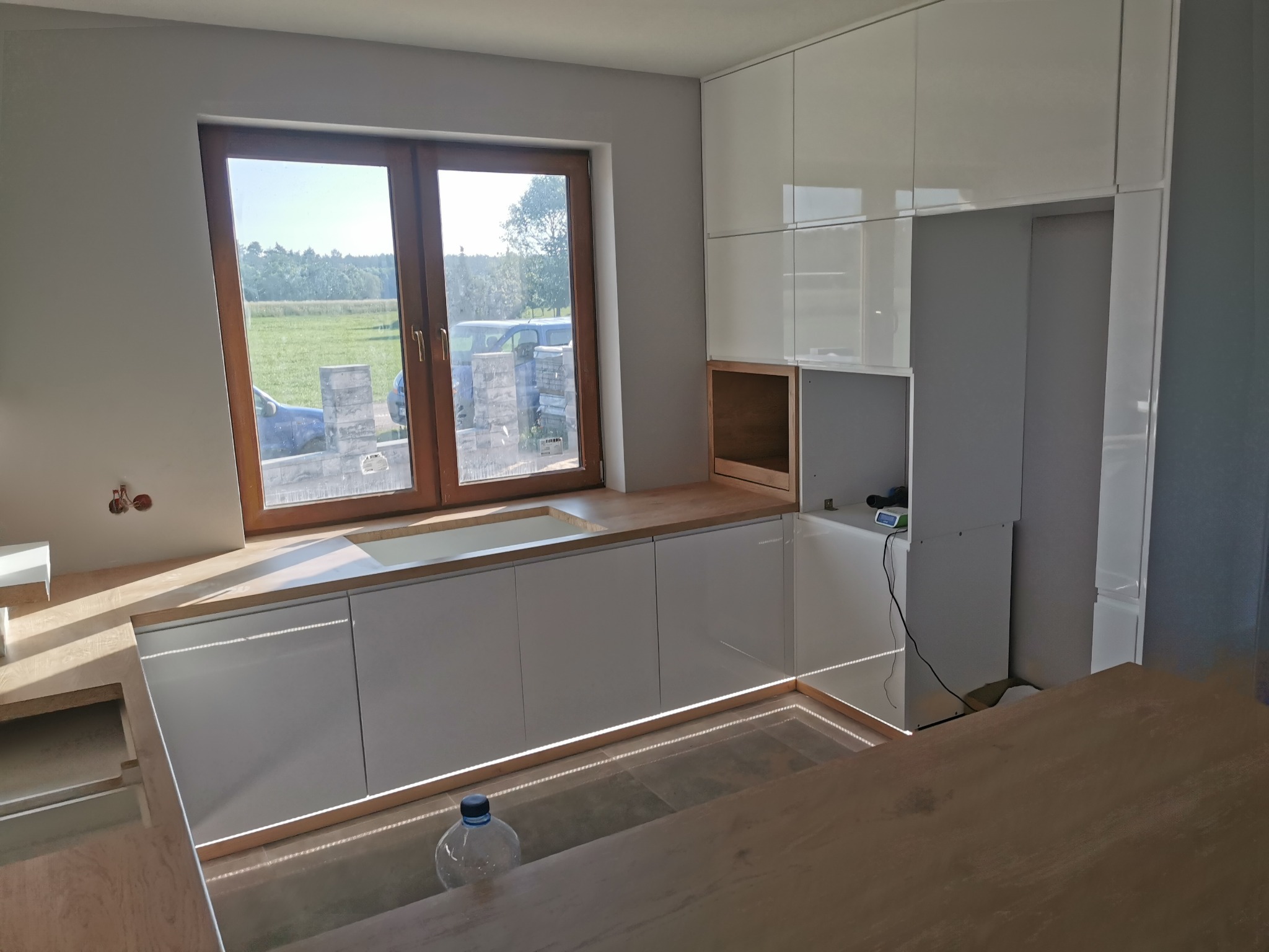 Nowoczesna kuchnia z białymi, lakierowanymi szafkami z podświetleniem LED, drewnianym blatem i widokiem na zewnątrz przez okno, częściowo zdemontowana, z butelką wody na blacie.