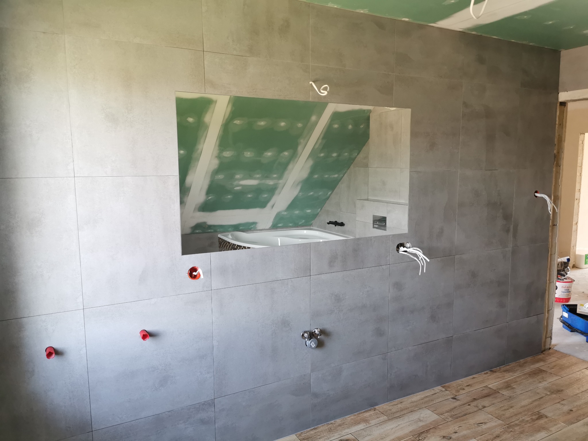 Szara łazienka w trakcie remontu z widocznymi płytkami na ścianie, przygotowanymi instalacjami wodnymi i elektrycznymi oraz częściowo ułożoną podłogą z paneli imitujących drewno. W lustrze odbija...