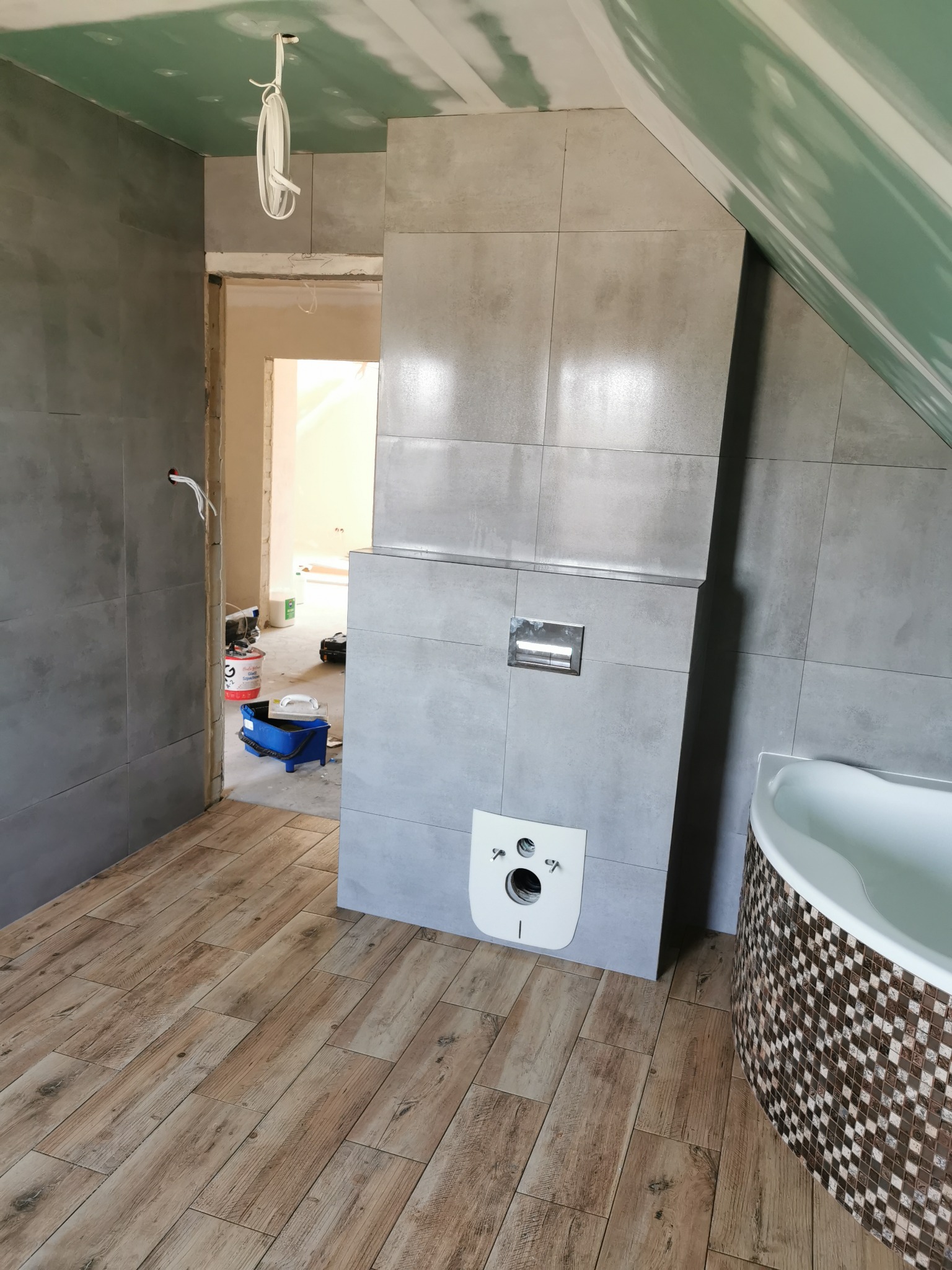 Wnętrze łazienki w trakcie remontu: szare płytki na ścianach, drewnopodobna podłoga, częściowo zabudowana toaleta podtynkowa, wanna obłożona mozaiką, widoczne narzędzia i materiały budowlane.