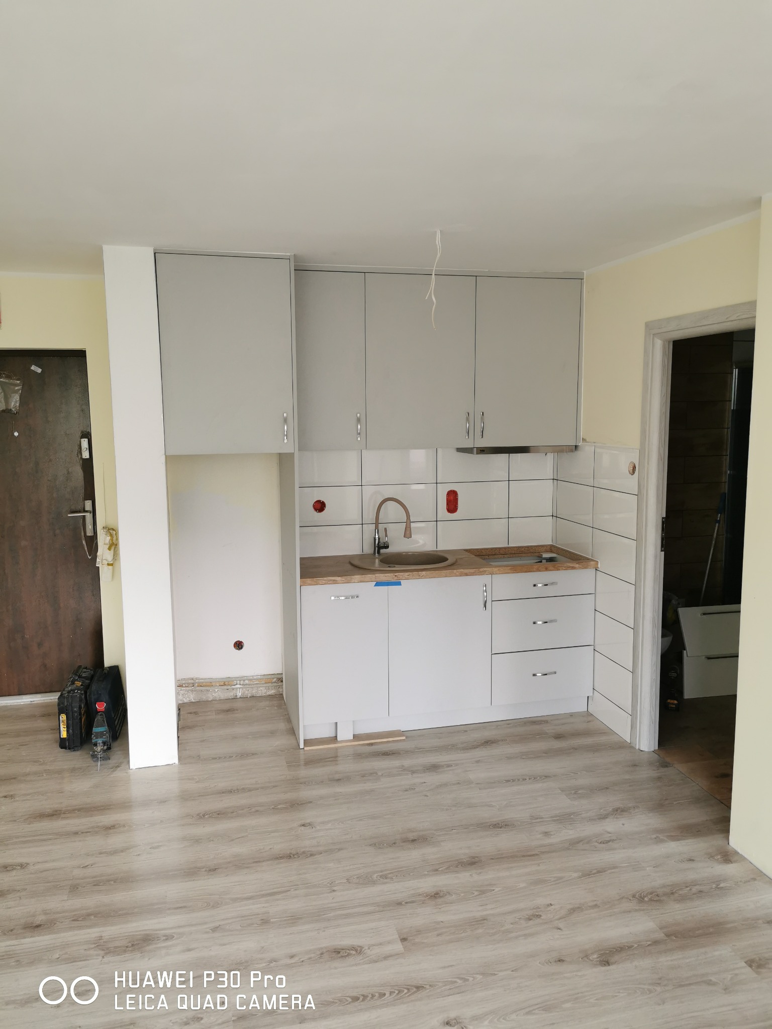 Jasnoszara zabudowa kuchenna z drewnianym blatem i brązową armaturą, białe kafelki na ścianie, widoczne narzędzia na podłodze przy drzwiach wejściowych.