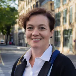 Portret uśmiechniętej kobiety w białej koszuli i czarnej marynarce na tle miejskiej ulicy z budynkami o jasnej elewacji.