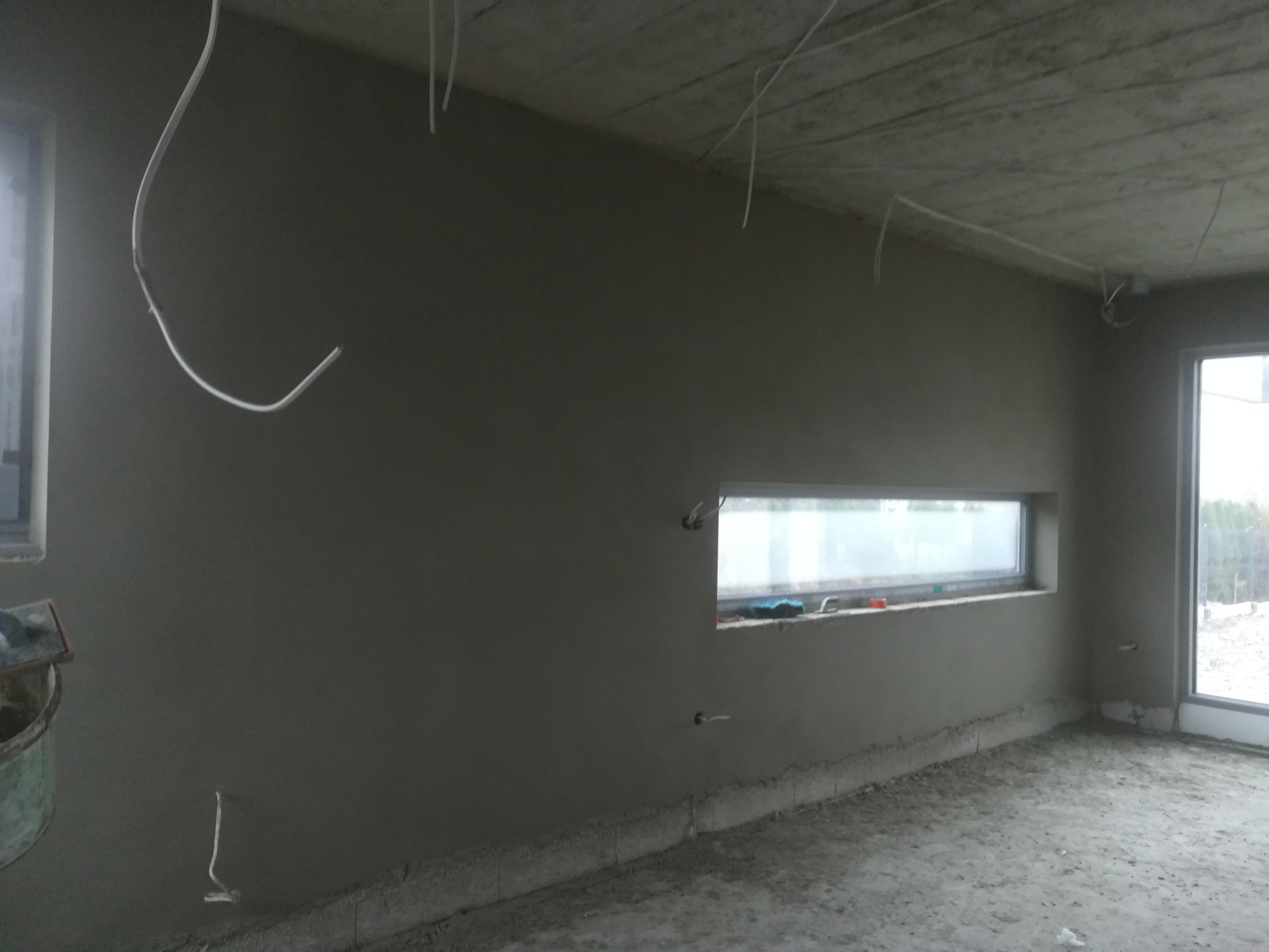 Wnętrze w stanie surowym po nałożeniu tynku gipsowego na ścianach, widoczne wystające przewody elektryczne, wąskie okno i wiadro z narzędziami malarskimi w rogu pomieszczenia.