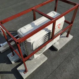 Biała jednostka zewnętrzna klimatyzacji marki Rotenso, zamontowana na betonowych podstawach i zabezpieczona czerwoną, metalową konstrukcją na czarnym, płaskim dachu.