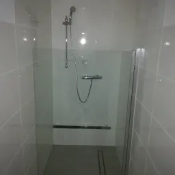 Nowoczesna kabina prysznicowa ze szklanymi drzwiami i odpływem liniowym w podłodze, wykończona białymi, błyszczącymi płytkami ściennymi z delikatną fugą.