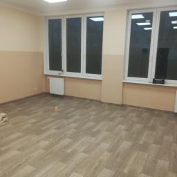 TomDom - Pusty pokój po remoncie z panelami podłogowymi w odcieniach brązu, pomalowanymi ścianami do połowy wysokości na kolor beżowy, z trzema oknami i białymi ramami oraz grzejnikiem pod jednym z nich...