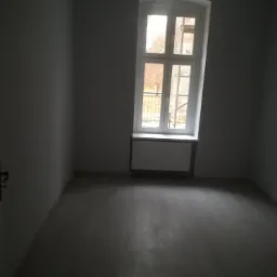 Pusty pokój z szarymi ścianami po świeżo położonych gładziach, widoczne duże okno z białymi ramami i kaloryfer pod nim.