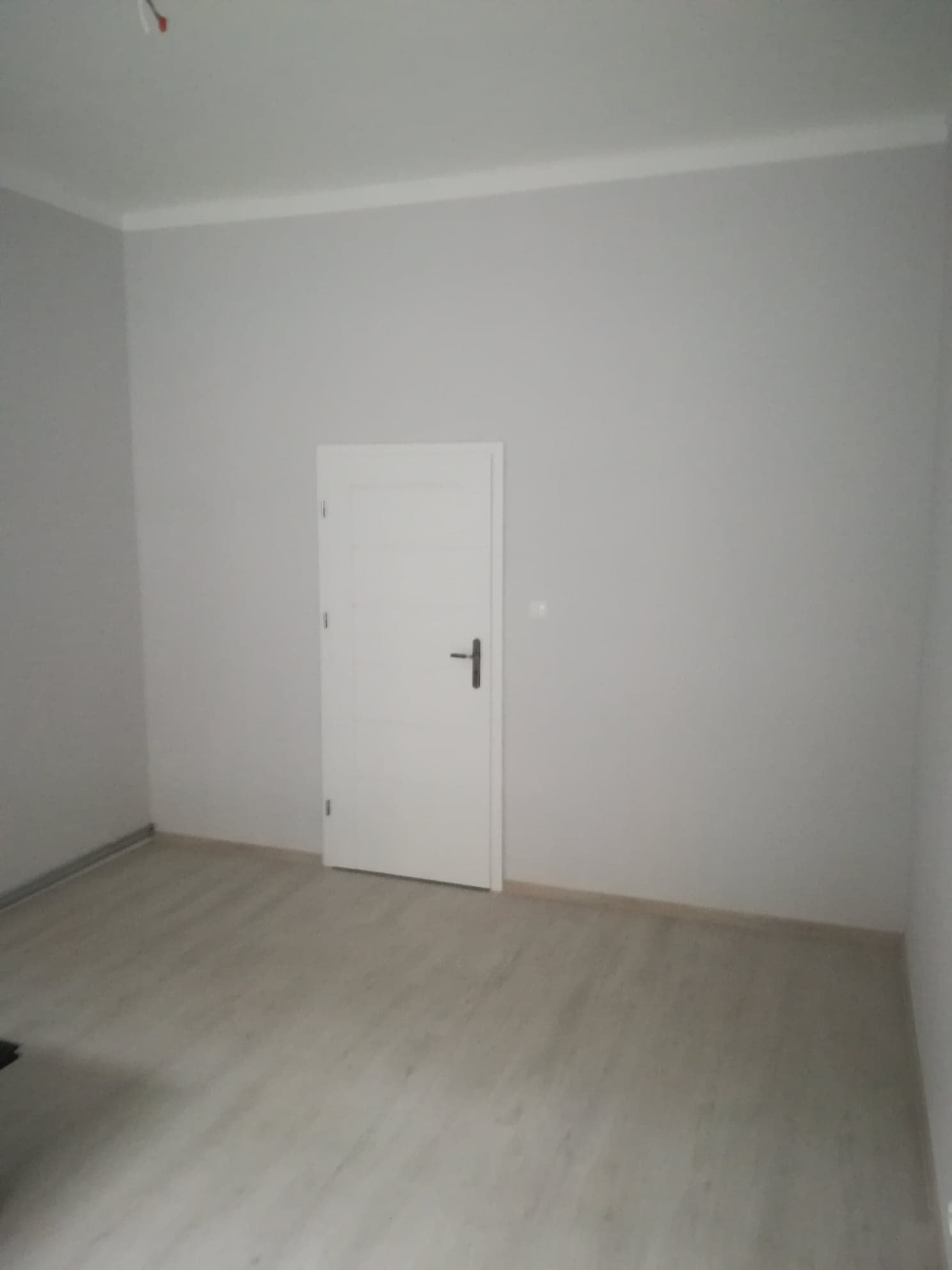 Pusty pokój po odświeżeniu: świeżo pomalowane szare ściany, białe listwy przypodłogowe i sufitowe, jasna drewnopodobna podłoga, białe drzwi wewnętrzne z czarną klamką.