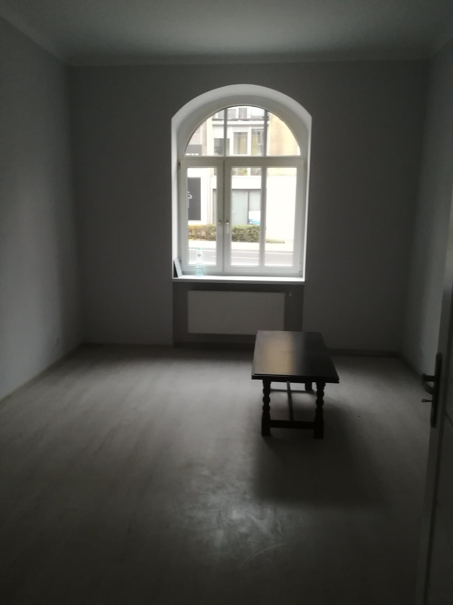 Pusty, wysprzątany pokój z szarymi ścianami i podłogą, z ciemnym stolikiem na środku i oknem łukowym z widokiem na budynek.