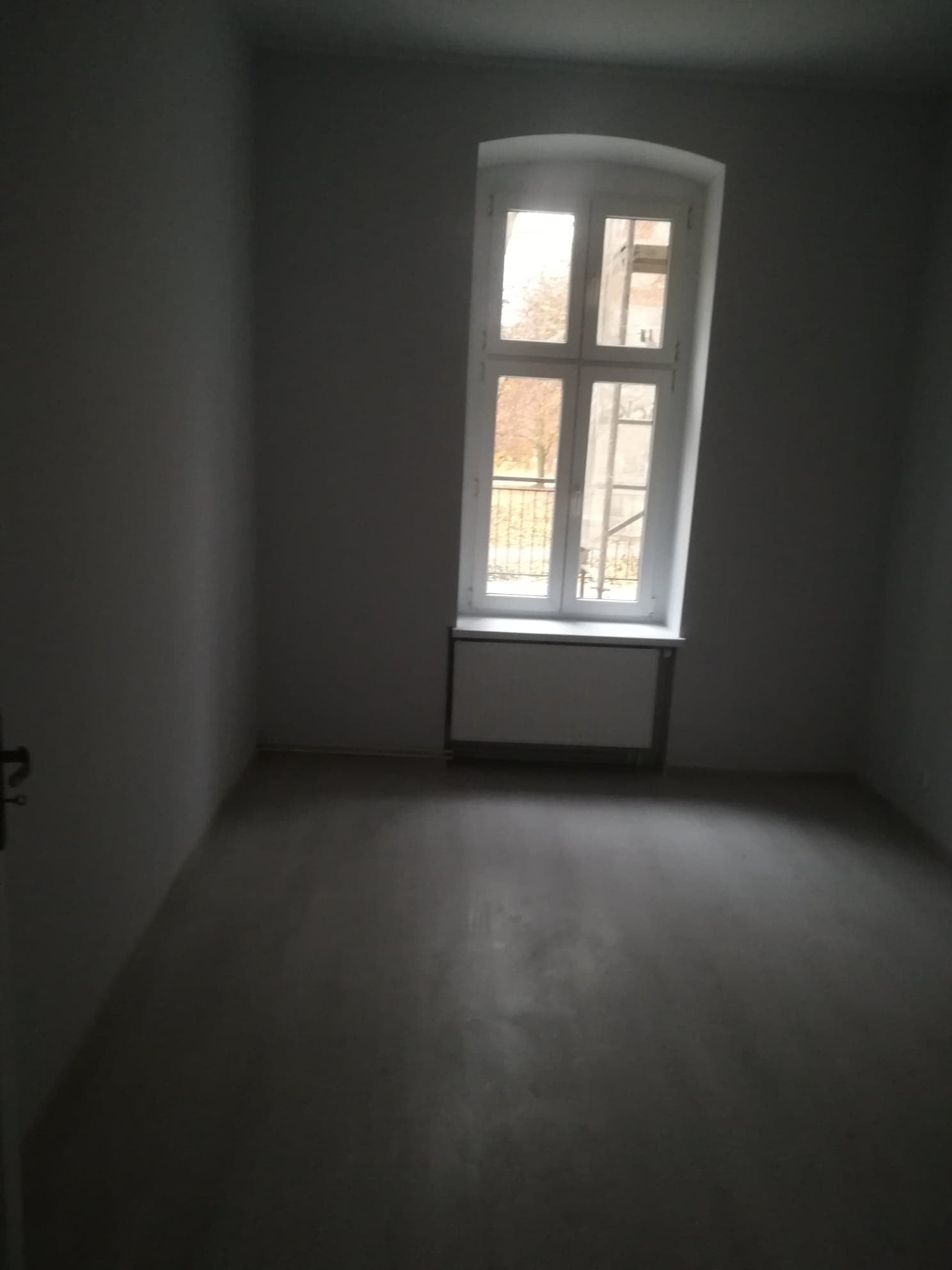 Pusty pokój z szarymi ścianami po świeżo położonych gładziach, widoczne duże okno z białymi ramami i kaloryfer pod nim.