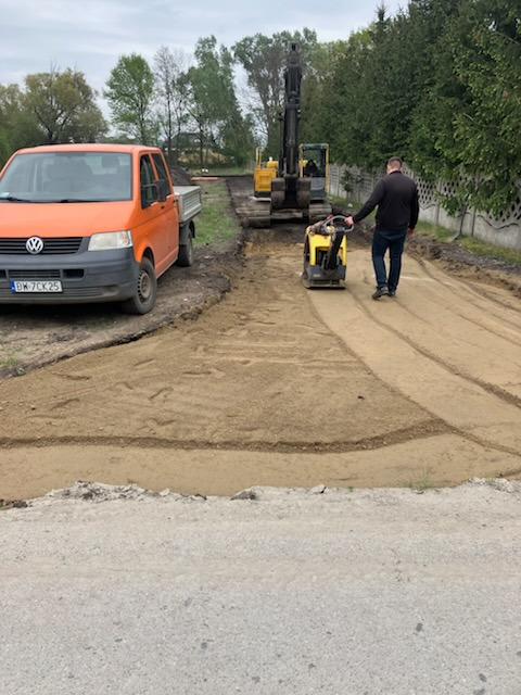 Utwardzanie drogi: operator zagęszcza piasek ubijarką, w tle koparka i pomarańczowy bus VW. Ślady ubijania widoczne na piasku. Dzień, pochmurno.