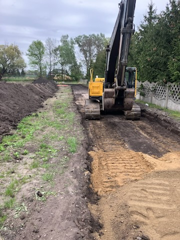 Żółta koparka gąsienicowa wyrównuje teren pod budowę drogi, widoczne ślady gąsienic na piasku, świeżo wykopana ziemia po bokach, ogrodzenie z prawej strony.