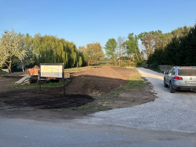 Teren po niwelacji z utwardzoną drogą dojazdową z białego gruzu. Na pierwszym planie tablica informacyjna projektu. W tle drzewa i zaparkowany samochód.