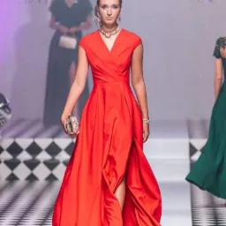 Modelka prezentuje długą czerwoną suknię z rozcięciem na wybiegu, z torebką w dłoni i biżuterią, na tle innych modelek i dekoracji.