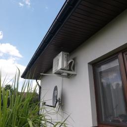 D-VENT Daniel Prusiński - Biała jednostka zewnętrzna klimatyzatora zamontowana na elewacji domu obok brązowego okna, widok z dołu na tle błękitnego nieba z chmurami.