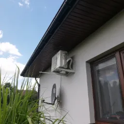 Biała jednostka zewnętrzna klimatyzatora zamontowana na elewacji domu obok brązowego okna, widok z dołu na tle błękitnego nieba z chmurami.