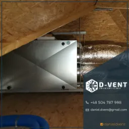 Srebrna centrala wentylacyjna zamontowana na poddaszu, widoczne rury wentylacyjne w izolacji aluminiowej i fragment niebieskiego przewodu elastycznego.