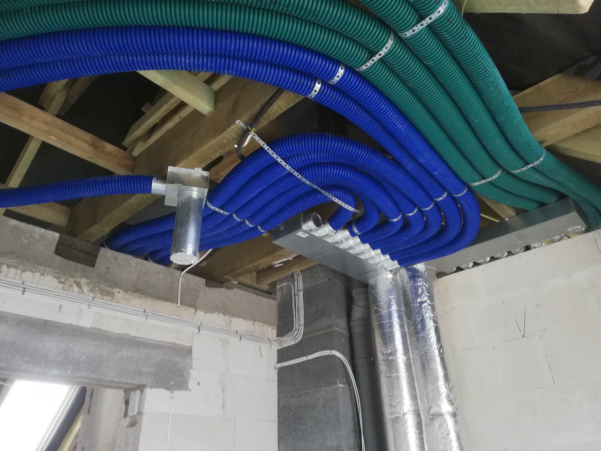 Niebieskie i zielone karbowane rury wentylacyjne przymocowane do drewnianej konstrukcji dachu w surowym wnętrzu budynku, z widocznymi elementami instalacji i przewodami.