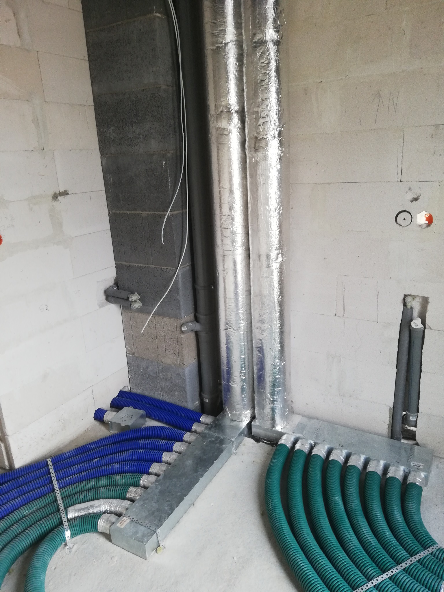 System kanałów wentylacyjnych z rurami w kolorze niebieskim i zielonym, podłączonych do skrzynki rozdzielczej w surowym wnętrzu budynku z betonowymi ścianami.