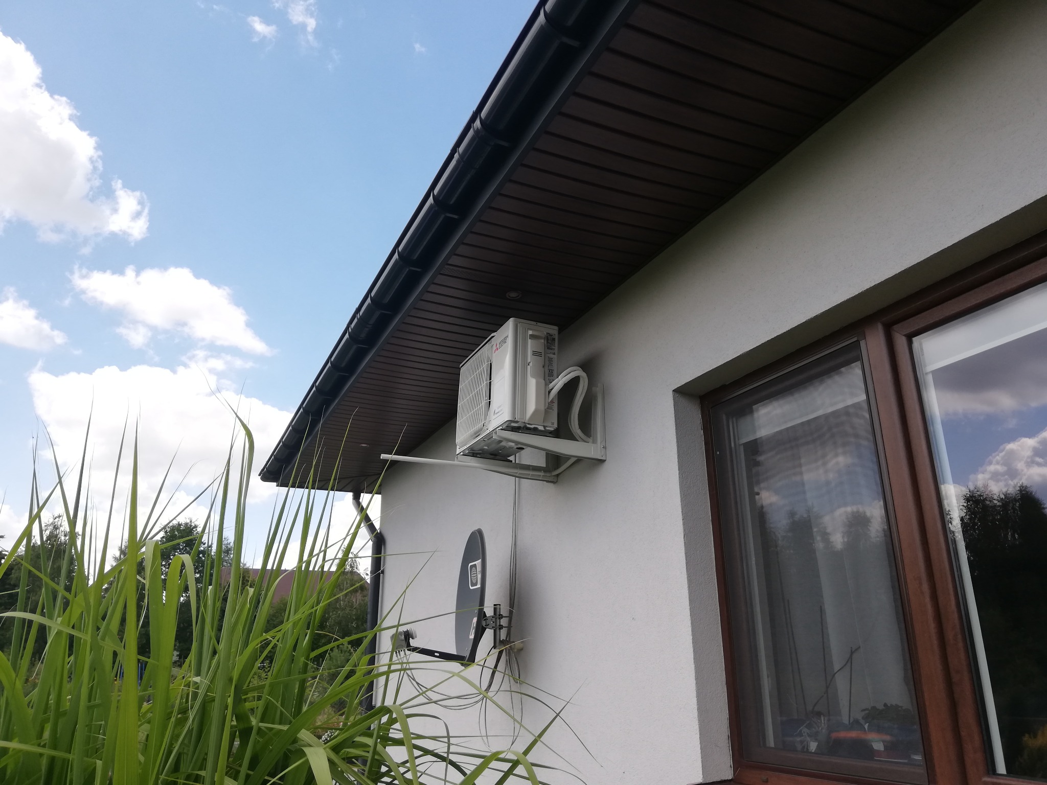 Biała jednostka zewnętrzna klimatyzatora zamontowana na elewacji domu obok brązowego okna, widok z dołu na tle błękitnego nieba z chmurami.